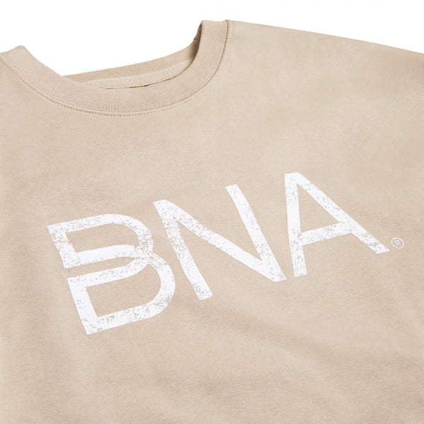 Detail shot of light beige crewneck sweatshirt with white distressed BNA logo centered on the chest.  BNA logo features a unique infinity "B."